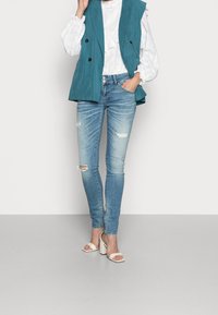 Blazer sans manches turquoise porté sur un chemisier blanc texturé, associé à un jean skinny bleu clair déchiré et des sandales blanches à talons.