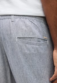 Nahaufnahme einer hellgrauen, strukturierten Hosentasche und eines Bunds, mit einem Teil eines weißen Hemdes und eines entspannten Arms einer Person sichtbar.