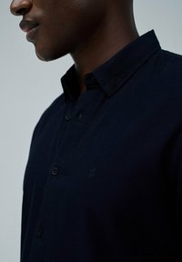 Camisa de botones azul marino con una tela texturizada, un pequeño detalle de logo y un cuello clásico. Presenta botones negros a lo largo de la tapeta.