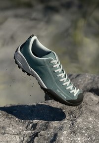 Világoskék túracipő, texturált felsőrésszel, szürke gumitömítésekkel és masszív talppal. A fűzők hangsúlyosan láthatóak.