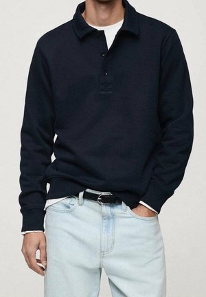 Camisa polo de manga larga azul marino hecha de algodón, con cuello, tapeta de tres botones y puños acanalados, llevada sobre vaqueros de mezclilla claros.