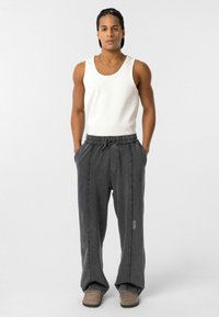 Grijze joggingsbroek met een trekkoord in de taille, zijnaad en logo-detail, gecombineerd met een witte tanktop en beige schoenen, tegen een eenvoudige achtergrond.