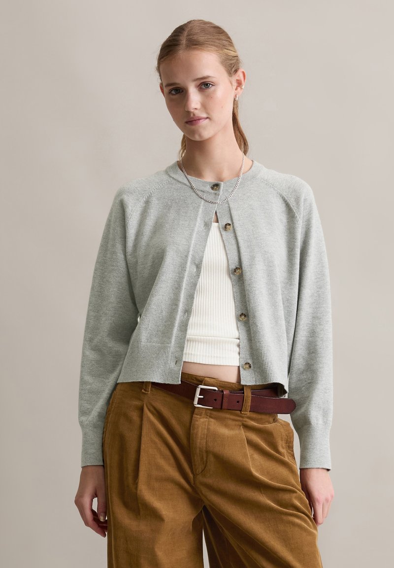 Cardigan grigio chiaro con scollatura rotonda, chiusura con bottoni e orlo a costine, indossato sopra una maglietta bianca a costine e abbinato a pantaloni di corduroy marroni.