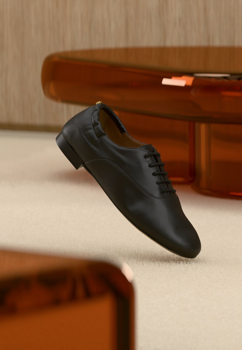 Chaussure habillée en cuir noir avec lacets posée sur un tapis beige, avec un mobilier marron translucide en arrière-plan.