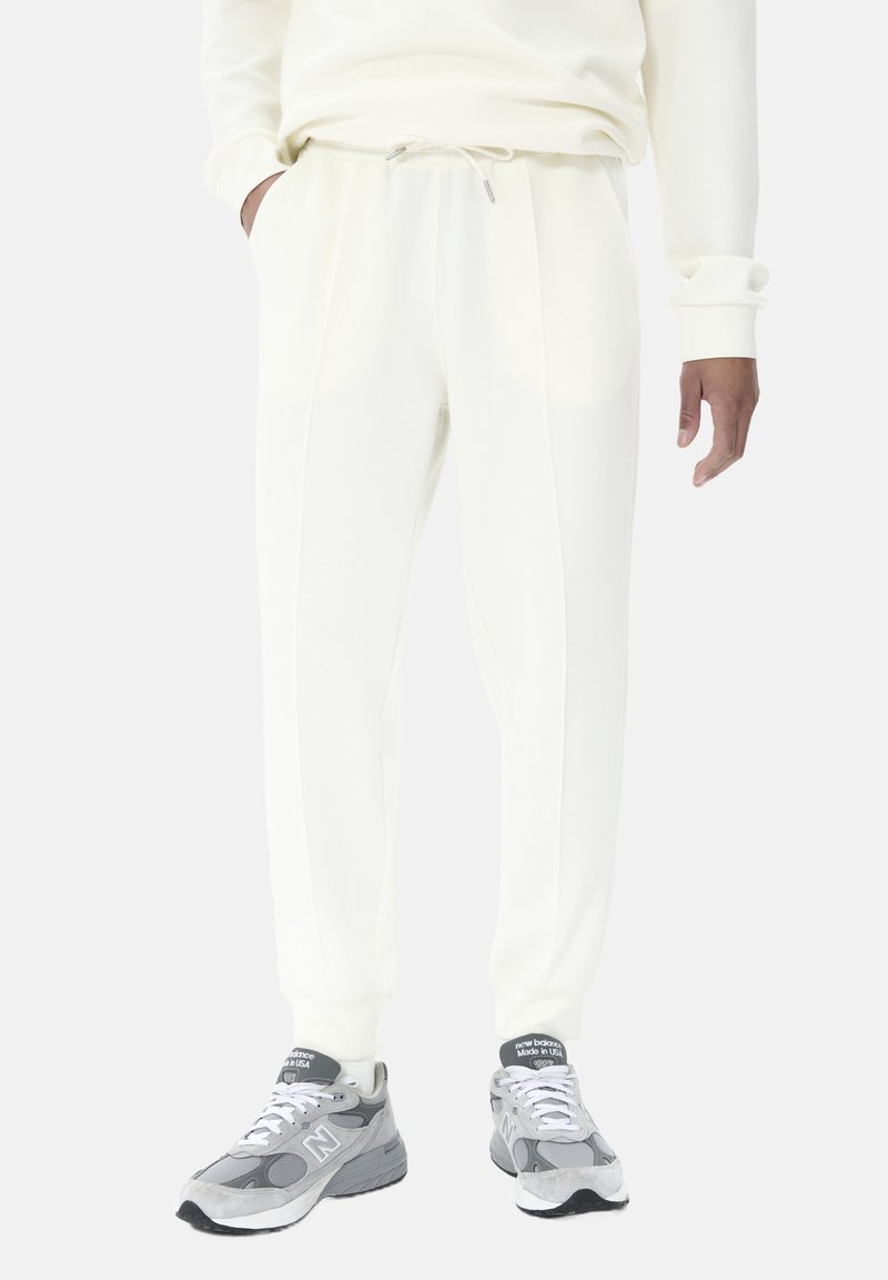 Hvide sweatpants med elastisk talje og snøre, der har sidelommer og en glat, blød tekstur. Kombineret med grå sneakers.
