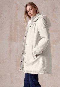 Lichtgrijze oversized jas met een pluizige, textuurrijke witte voorkant, drukknoopsluiting en een capuchon. Heeft zijzakken en elastiche manchetten.