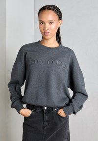 Felpa grigia con scollo a girocollo e testo ricamato "Marc O’Polo". Abbinata a una gonna nera, mani nelle tasche, design casual.