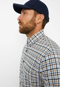 Karierte Hemdbluse mit blauen, braunen und weißen Mustern; besitzt einen strukturierten Kragen und dunkle Knöpfe. Kombiniert mit einer marineblauen Kappe.