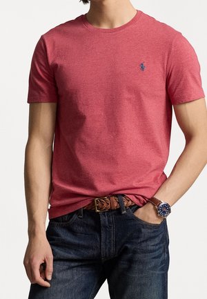 Rode katoenen t-shirt met een ronde hals en korte mouwen, met een klein blauw logo op de borst. Draagt met denim jeans en een gevlochten riem.