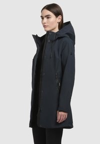 Marinblå vattentät parka med huva, frammensknapp, knäppbar klaff och justerbara ärmslut. Har sidfickor och en strömlinjeformad silhuett.