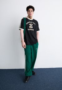 Melna Adidas t-kreka ar zelta logotipu, zaļas Adidas sporta bikses un melnas kedas. Apģērbs ir ar trim baltām svītrām uz biksēm.