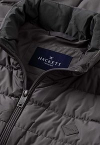 Donkergrijze pufferjas met een ritssluiting aan de voorkant, voorzien van een logolabel aan de binnenkant en een doorgestikte structuur. Glad stof met een hoge kraag.