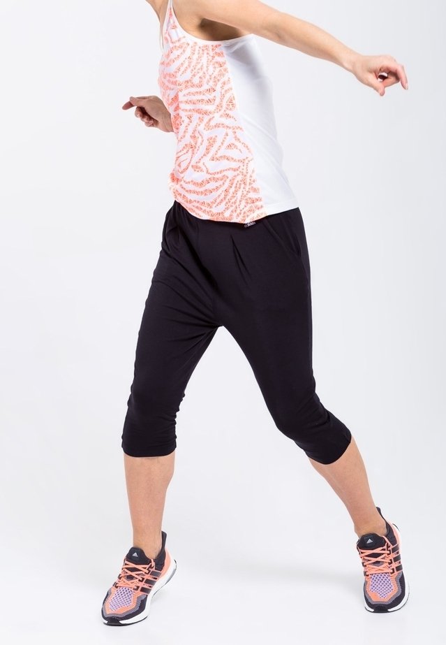 Personne étirant les jambes, portant un pantalon noir court, un débardeur blanc avec motif orange, et des chaussures de course avec des accents roses sur fond blanc.