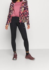 Fialová bunda s květinovým vzorem, růžový uzlový top, černé legíny a hnědé sportovní boty. Mezi charakteristické rysy patří kapuce a zapínání na zip.