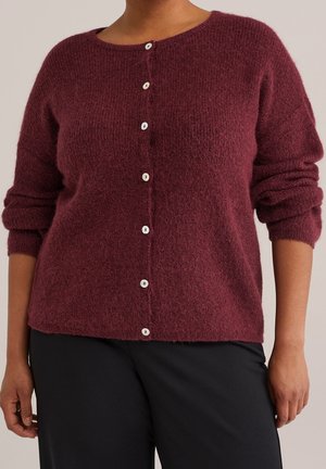 Femme portant un cardigan bordeaux en laine à manches longues boutonné, avec les manches retroussées, et un pantalon noir, sur un fond uni.