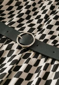 Tissu à motifs noir et blanc avec une ceinture en cuir vert ornée d'une boucle circulaire argentée, mettant en valeur la texture et le contraste.