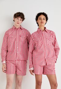 Rot-weiße karierte Hemden und Shorts mit klassischem Gingham-Muster, Knopfleiste und Brusttaschen, präsentiert von zwei Models.