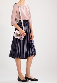 Donna che indossa una blusa rosa pallido, una gonna midi plissettata in velluto blu navy, ballerine nere con perle e una piccola borsa a tracolla floreale rosa.