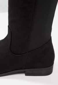 Gros plan d'une botte cheville en suède noir avec un petit talon et un panneau latéral élastique sur un fond blanc.