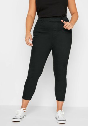 Vrouw draagt zwarte hoge taille cropped broek, zwart mouwloos topje en witte sneakers, staand tegen een eenvoudige achtergrond.