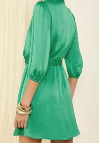 Vihreä satiinimekko, jossa on muotoiltu vyötärö, kolmannen neljänneksen puffihihat ja sileä pinta. Mekossa on yhteensopiva vyö ja virtaava hame.
