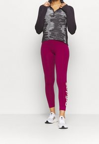 Pitkähihainen, vetoketjullinen urheilupaita, jossa on harmaa ja musta kuvio, yhdistettynä tiukkoihin kirkkaanpinkkeihin legginsseihin, joissa on valkoinen "adidas"-logo.
