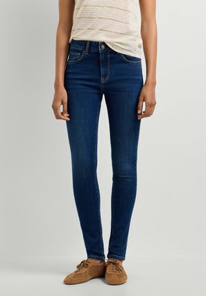 Persoon die donkerblauwe skinny jeans en bruine vetersneakers draagt, staat met de handen ontspannen langs het lichaam tegen een effen achtergrond.