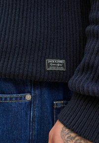 Navy ribbed knit sweater with a black logo patch from Jack & Jones, paired with blue denim jeans featuring yellow stitching.