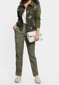 Olivgrüne Jeansjacke mit floralem Stickerei und Knopfverschluss, kombiniert mit olivfarbenen Cargo-Hosen mit Farbspritzern und Fronttaschen.