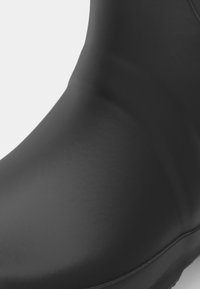 Botte noire imperméable avec une texture lisse et mate, une forme courbée et une couture renforcée sur le bord. Pas de quincaillerie ou de motifs visibles.