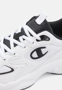 Valkoinen urheilukenkä, jossa verkkokangasta ja synteettisiä materiaaleja, mustat yksityiskohdat, valkoiset nauhat ja ommeltu logo sivulla.