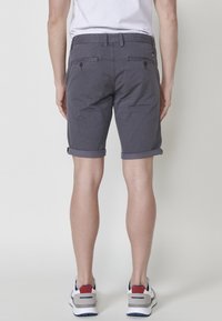 Muž v tmavošedých šortkách s vyhrnutými nohavicami, bielych teniskách s červenými a modrými akcentmi a bielom tričku, stojí proti svetlému pozadiu.