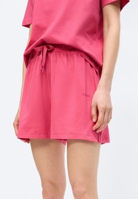 Femme portant un short rose vif à taille élastique avec cordon de serrage, ourlet court, et un logo "BOSS" discret sur la cuisse gauche.