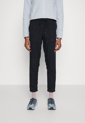 Persoon die zwarte broek met trekkoord, lichtblauw jack met rits, roze sokken en blauwe sportschoenen draagt, staand tegen een witte achtergrond.
