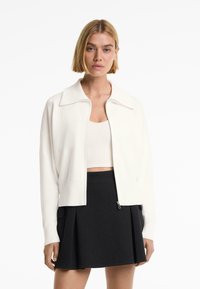 White zip-up cardigan with a wide collar, ribbed cuffs, and hem. Paired with a cropped beige top and black pleated skirt.
