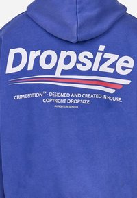 Niebieska bluza z kapturem z dużym, białym napisem "Dropsize", czerwonymi i białymi paskami oraz napisem "Crime Edition" poniżej. Miękki materiał z kapturem.