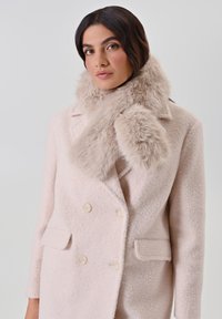 Manteau en laine rose pâle avec des boutons croisés et deux poches latérales, complété par une douce écharpe en fausse fourrure beige autour du cou.