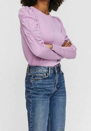 Osoba ze skrzyżowanymi ramionami, ubrana w lawendowy top z długim rękawem z marszczonymi rękawami oraz wysokim stanem niebieskich dżinsów, na jednolitym tle.
