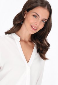 Biała bluzka typu v-neck z kołnierzykiem, gładka faktura i długie rękawy. Modelka ma okrągłe czarne kolczyki. Włosy są faliste i sięgają ramion.