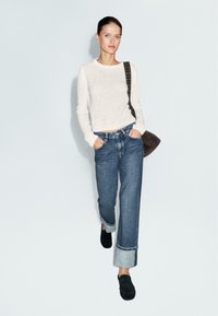 Donna che indossa un maglione bianco a maglia, jeans blu a vita alta con ampi polsini, scarpe nere e porta una borsa a tracolla marrone scuro, in piedi contro uno sfondo neutro.