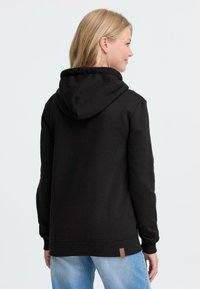 Hoodie noir avec une capuche spacieuse, en tissu doux, avec des poignets et un ourlet côtelés. Présente une petite étiquette de marque sur le côté inférieur.