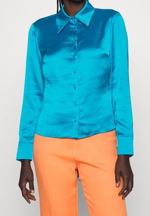Osoba ubrana w błyszczącą, turkusową koszulę z długim rękawem zapinaną na guziki, połączoną z pomarańczowymi spodniami o wysokim stanie i prostym kroju na tle jednolitego koloru.