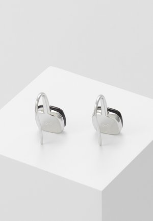 Boucles d'oreilles en métal argenté avec un design abstrait et courbé, ainsi qu'un petit détail gravé, exposées sur un socle cubique blanc.