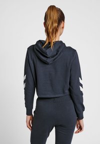Avskuren marinblå hoodie med dragsko i huvan och ribbade muddar. Har vita chevron-mönster på ärmarna. Mjuk, strukturerad textil.