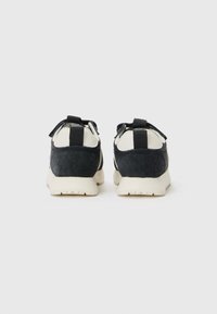 Baskets en suède et cuir noir avec des accents blancs, deux languettes à l'arrière et une semelle en caoutchouc texturée. Design simple avec des couleurs contrastées.