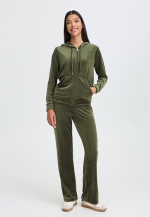 Vrouw staand, gekleed in een olijfgroene velours zip-up hoodie en bijpassende broek met witte sneakers, glimlachend tegen een effen achtergrond.
