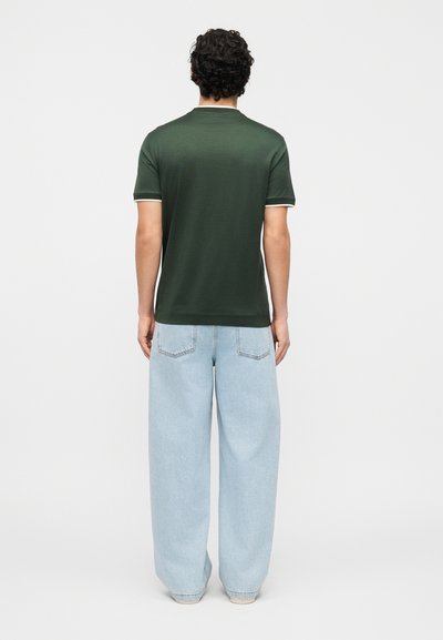 Άνδρας με σγουρά μαλλιά, στέκεται, φοράει σκούρο πράσινο κοντομάνικο πουκάμισο και άνετο ανοιχτό μπλε τζιν, μπροστά σε ένα απλό λευκό φόντο.