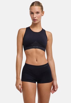 Vrouw draagt een zwarte sportbeha met hoge hals en bijpassende zwarte strakke shorts, staand tegen een witte achtergrond.