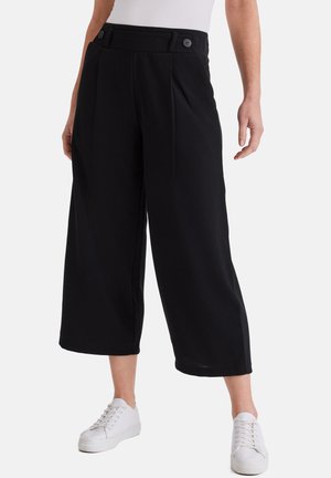 Frau trägt schwarze, hoch taillierte, weite, verkürzte Hosen mit Knopfdetails, kombiniert mit weißen Sneakern, stehend vor weißem Hintergrund.