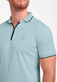 Pasaulio mėlynos polo marškiniai su užtrauktuko apykakle, juodu akcentu ir tekstūruotu audiniu. Priekyje yra mažas logotipas. Trumpomis rankovėmis.